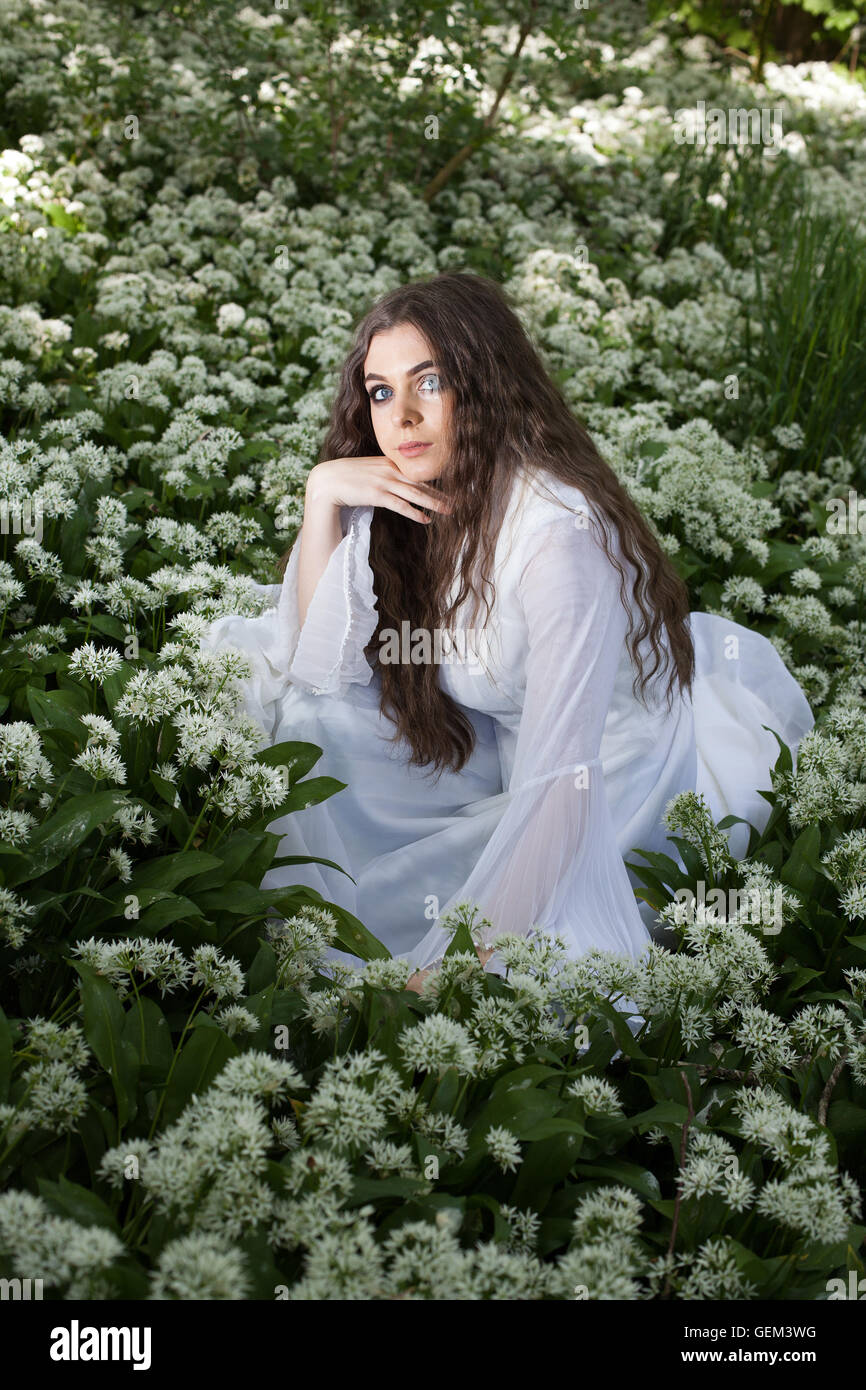 Schöne Frau in einem langen weißen Kleid sitzt im weißen Blüten auf einem Waldboden Stockfoto