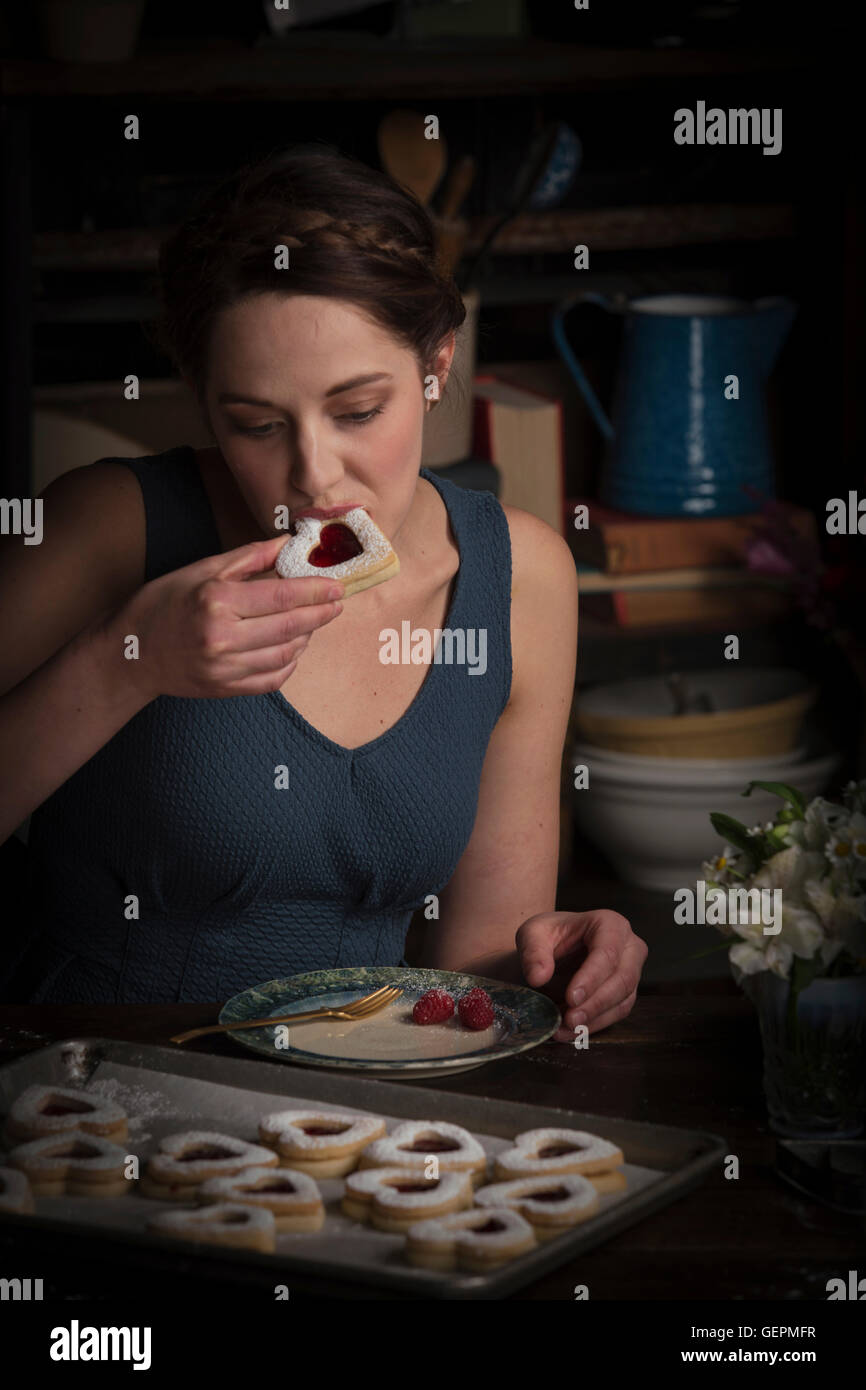 Valentinstag Backen, junge Frau sitzt in einer Küche Essen ein Herz geformt Keks. Stockfoto