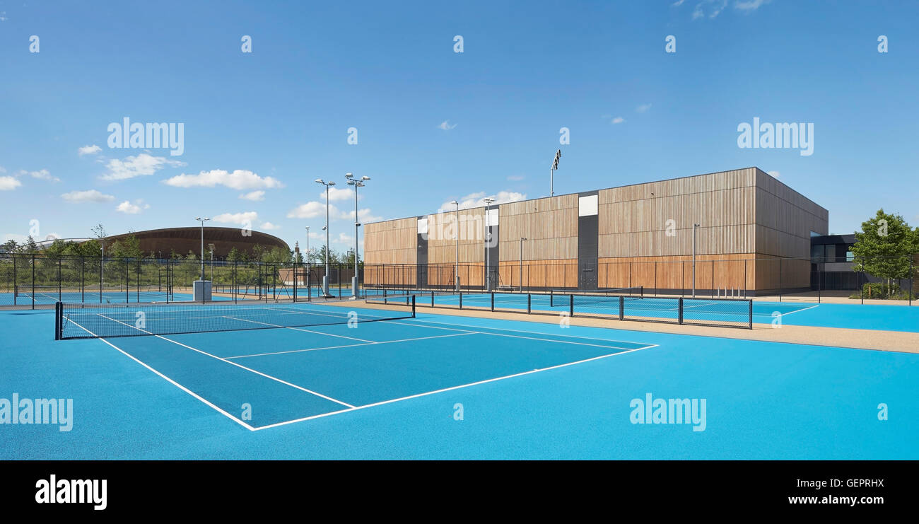 Helle blaue Tennisplatz mit Holz verkleidet Tenniscenter und Velodrom darüber hinaus. Eton Manor - Lee Valley Hockey und Tennis Centre, London, Vereinigtes Königreich. Architekt: Stanton Williams, 2014. Stockfoto