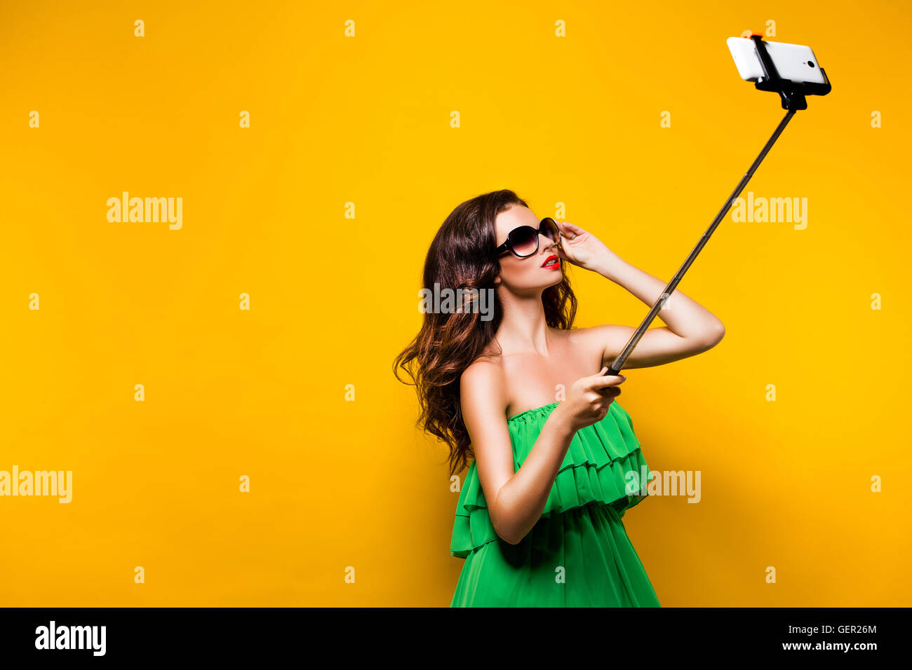 Porträt des jungen Modells im grünen Kleid und Sonnenbrille posiert dabei Selfie vor gelbem Hintergrund. Stockfoto