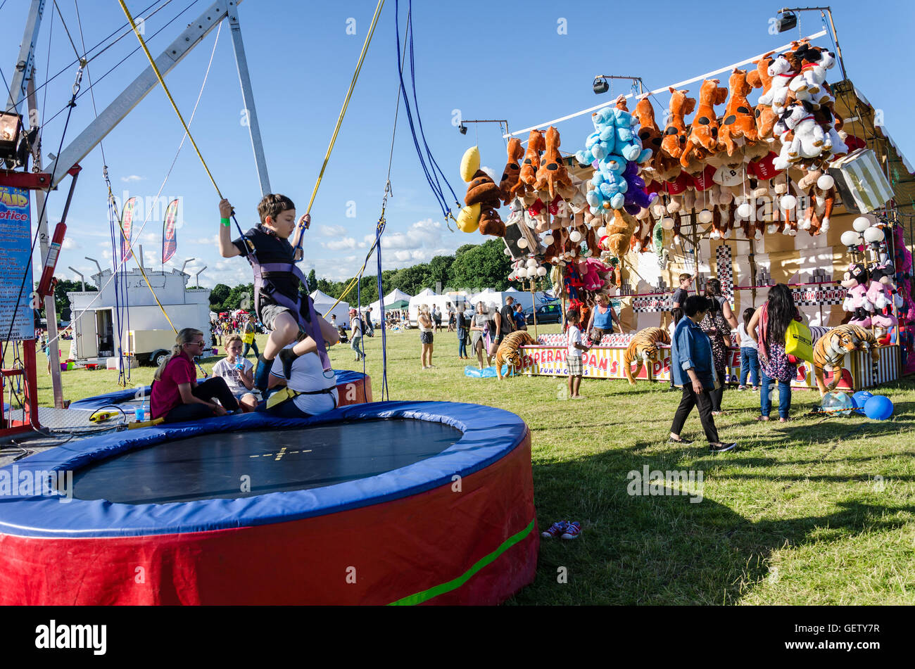 Ein kleiner Junge hat Spaß beim Springen auf einem Bungee-Trampolin auf einem Rummelplatz. Dahinter steckt eine "Tin Can Alley" Spiel mit jede Menge Preise. Stockfoto