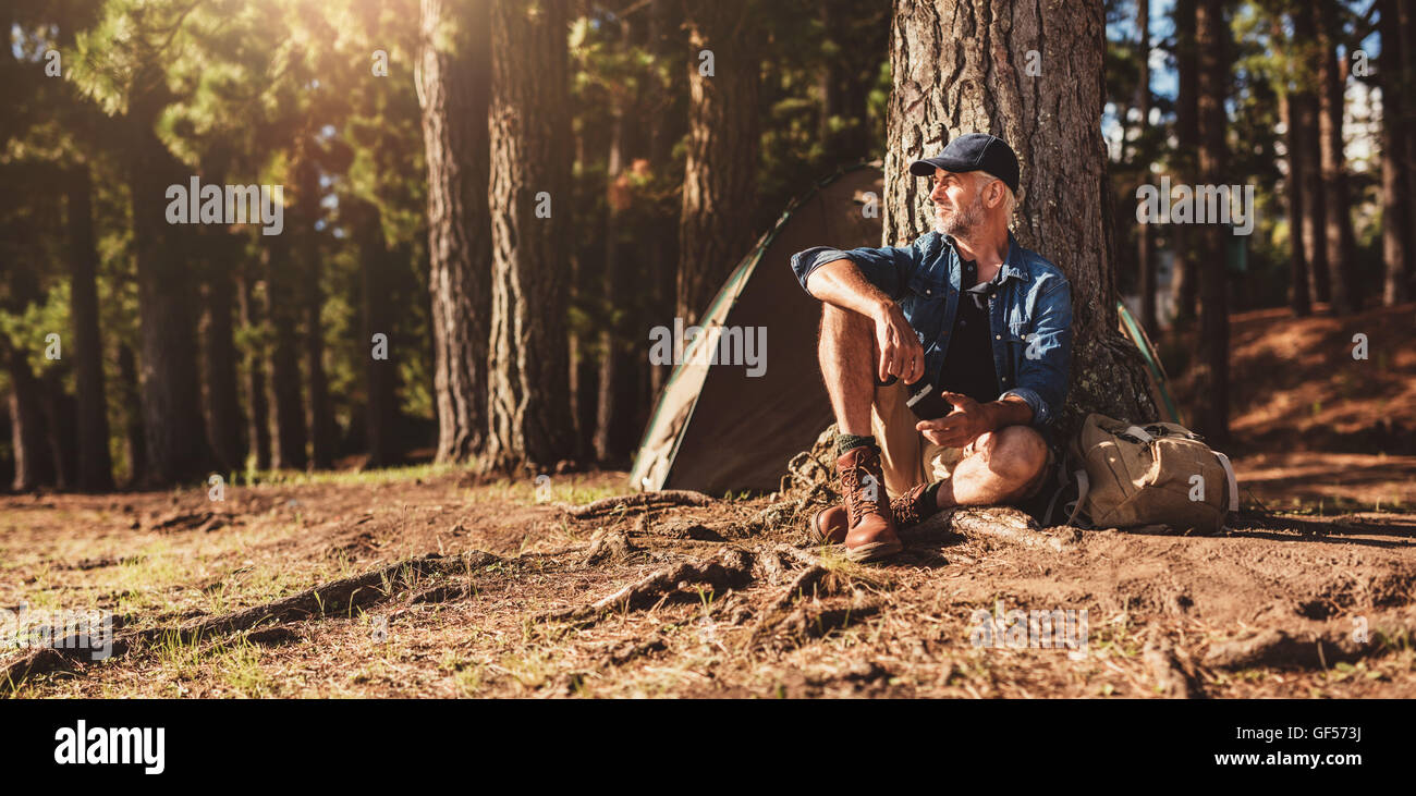 Porträt von senior Mann von einem Baum mit einem Zelt im Hintergrund sitzt. Reifer Mann sitzt auf einem Campingplatz. Stockfoto