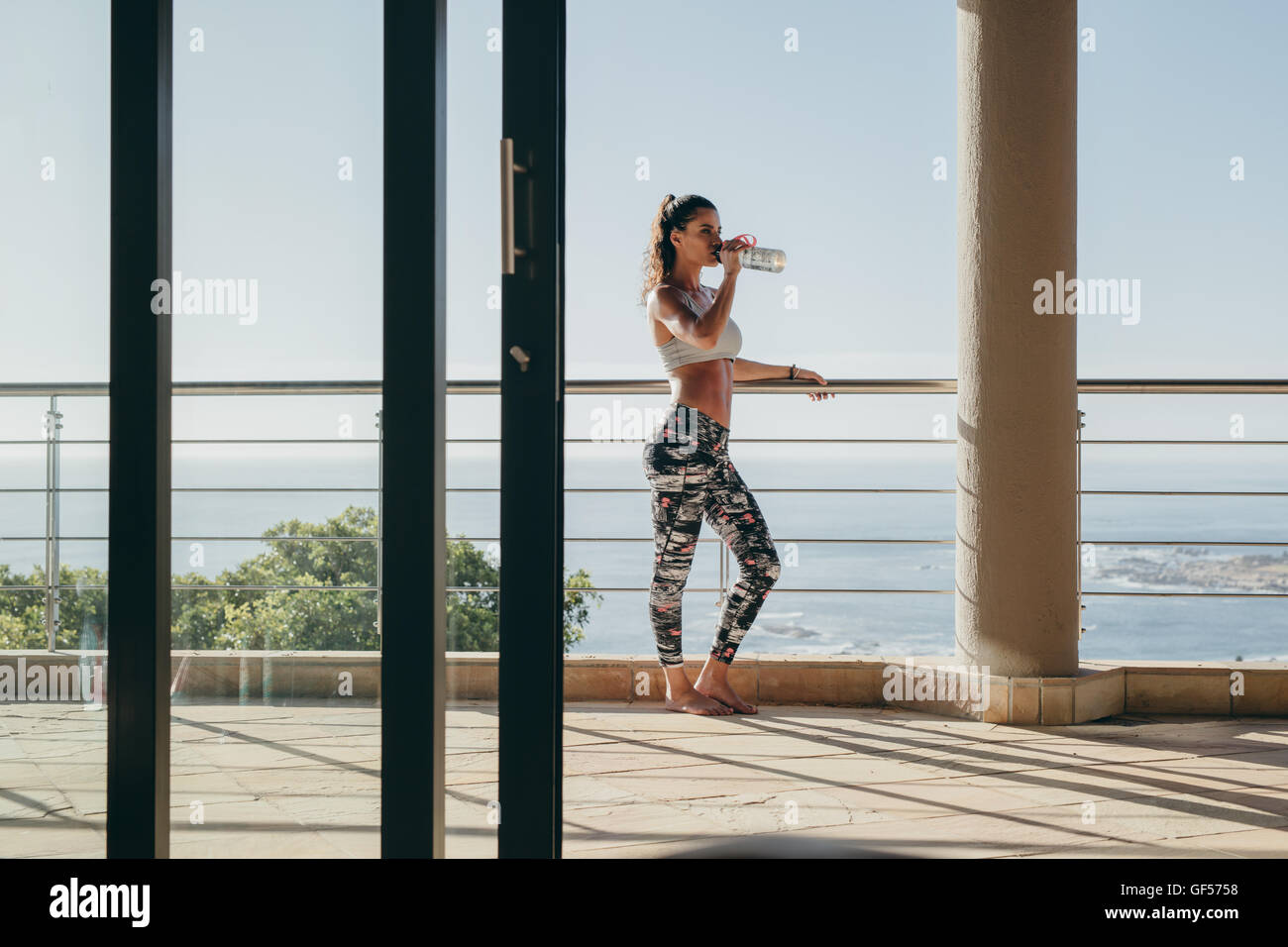 Außenaufnahme einer jungen Frau, die auf dem Balkon steht und Wasser aus der Flasche trinkt. Fitness-Frau macht Pause vom Training. Stockfoto