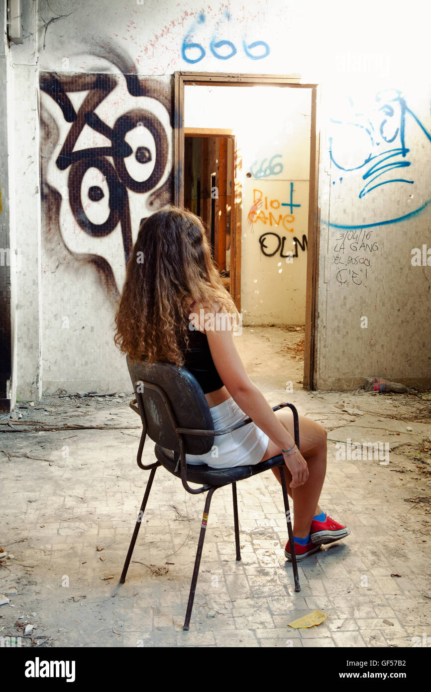 Junge Frau sitzt auf einem Bürostuhl in einen Flur von einem verlassenen psychiatrischen Krankenhaus mit dämonischen Schriften an den Wänden Stockfoto