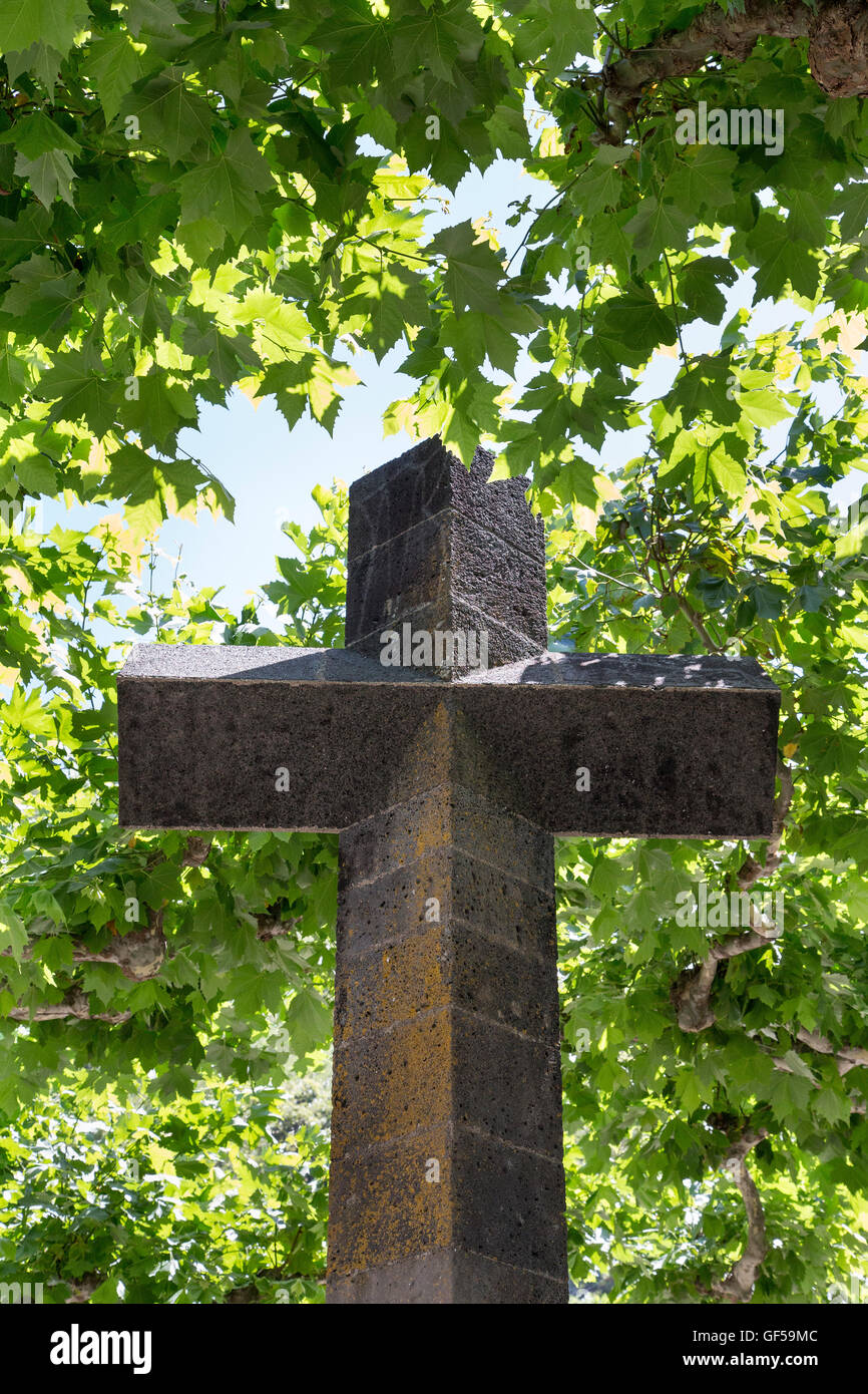 Katholisches Kreuz, aus vulkanischem Gestein, aus Basalt. Religiöses Wahrzeichen im Dorfzentrum von Lajes. Hintergrund gefüllt mit grünen Ahornblättern. Azoren, Pico. Stockfoto
