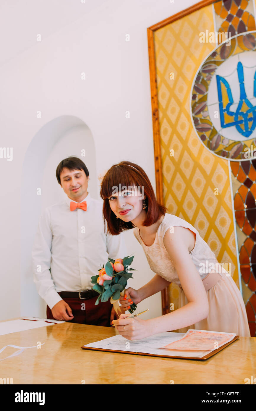 Braut, Heiratsurkunde oder Hochzeit Vertrag unterzeichnen und Blick in die Kamera, während der Bräutigam hinter Stockfoto