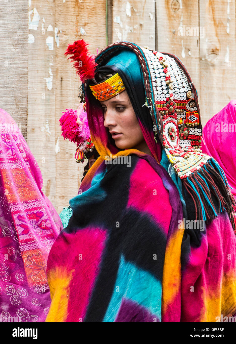 Kalash Frau trägt einen traditionellen Kauri Muschel Kopfschmuck und einem bunten Schal. Sie steht vor einem Haus Kalash. Stockfoto