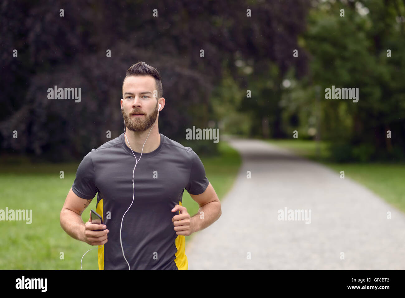 Fit bärtige junge Mann joggen durch den Park, das Anhören von Musik auf seinem Handy, Oberkörper, nähert sich die Kamera mit Kopie Stockfoto