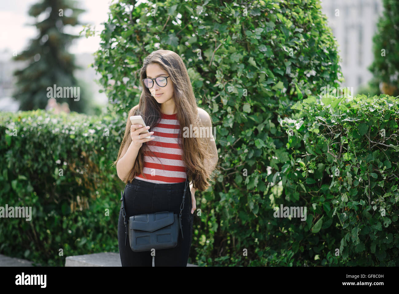 Junge asiatische Frau mit Brille mit Smartphone und Sms senden. Junge Frau auf Stadt-Hintergrund mit grünen Blättern. Stockfoto