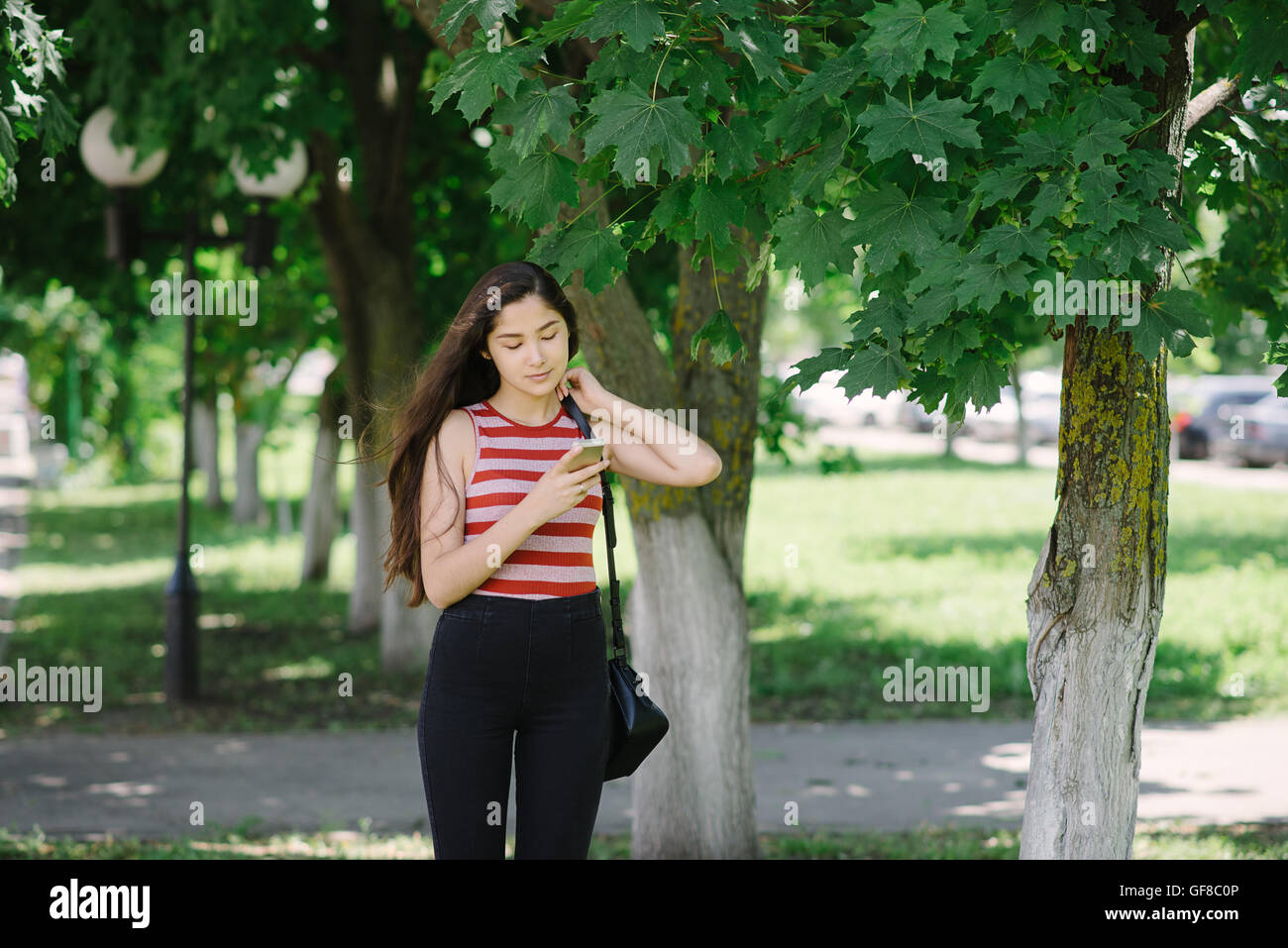 Junge Asiatin mit Smartphone und Sms senden. Junge Frau auf Stadt-Hintergrund mit grünen Blättern. Mädchen im Park. Stockfoto