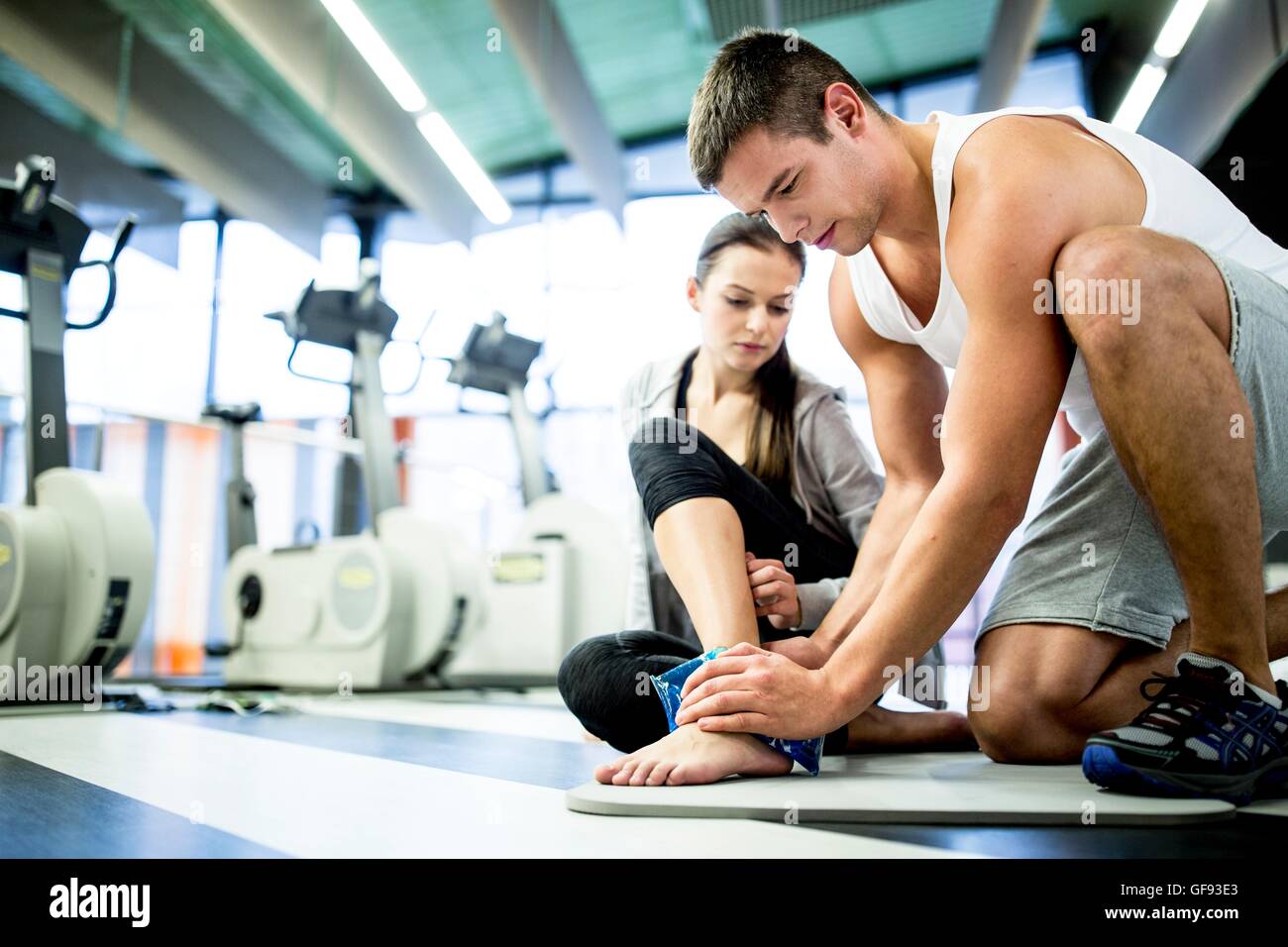 EIGENTUM FREIGEGEBEN. -MODELL VERÖFFENTLICHT. Junger Mann mit Eisbeutel auf junge Frau Knöchel im Fitness-Studio. Stockfoto