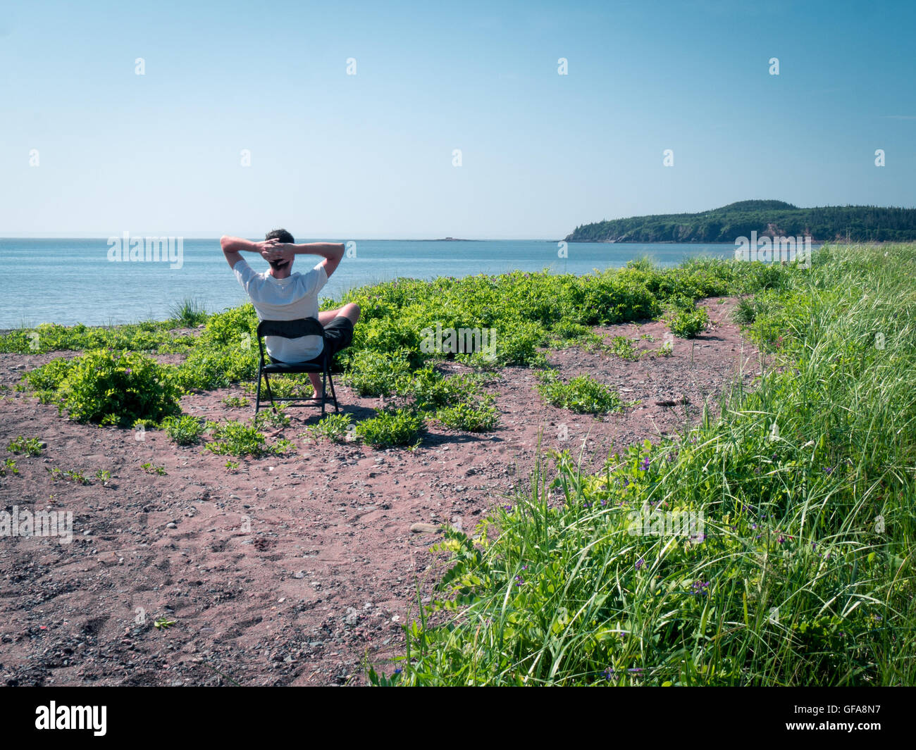 Mann sitzt auf einem Stuhl am steinigen Strand mit Blick auf eine Bucht mit Hügeln im Hintergrund Stockfoto