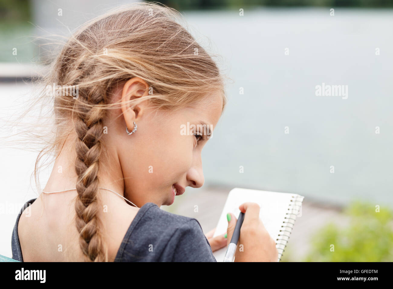 Junges Mädchen mit Zöpfen, die Skizze im Notizbuch sitzen in der Nähe von einem Teich Stockfoto