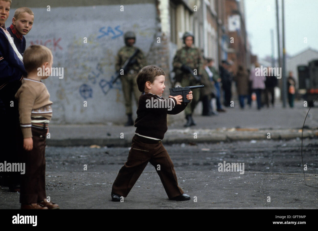 Junge, ein Kind, das mit einer Spielzeugpistole spielt, während der Probleme auf Passanten schießt. Belfast 1981 Nordirland 1980er Jahre Großbritannien. Britische Soldaten patrouillieren auf der Straße dahinter. Downtown City Shopping Street in Belfast. Danny Devine Boy mit Spielzeugpistole lebte hier in der Beechmount Avenue „RPG Avenue“, benannt nach den Raketenwerfer der IRA. HOMER SYKES Stockfoto