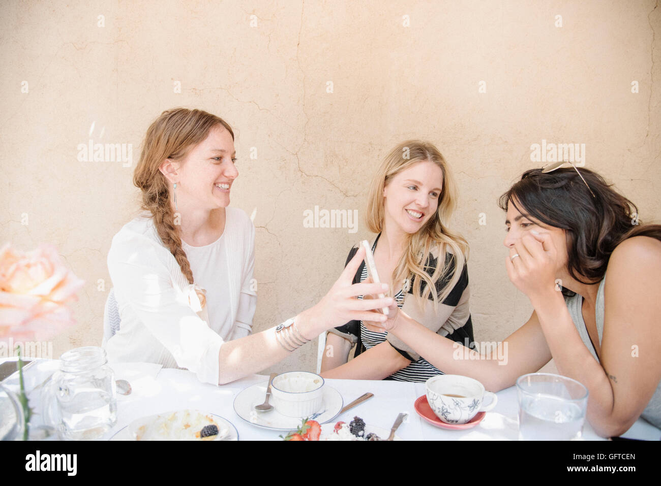Drei lächelnden junge Frauen sitzen um einen Tisch mit Speisen und Getränken, Blick auf ein Handy Stockfoto