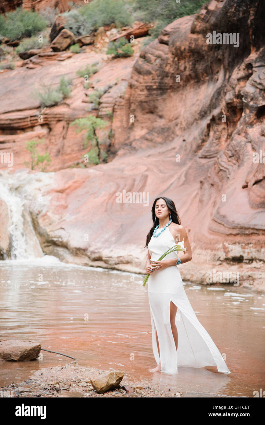 Junge Frau mit langen braunen Haaren, trägt ein langes weißes Kleid steht an einem Fluss halten Arum Lilies Stockfoto