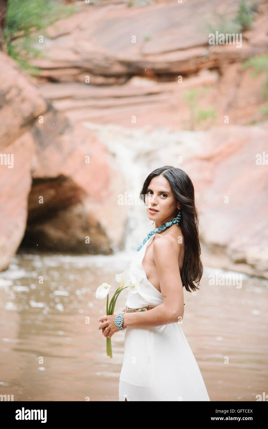 Junge Frau mit langen braunen Haaren, trägt ein langes weißes Kleid steht an einem Fluss halten Arum Lilies Stockfoto