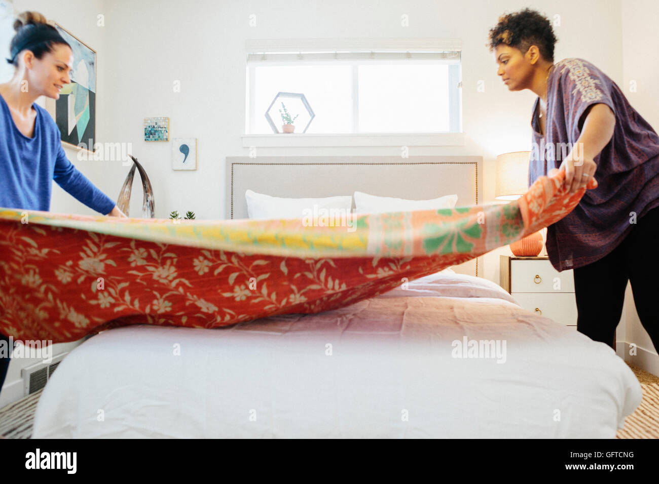 Zwei Frauen, aus denen ein Bett verbreiten eine gemusterte Decke über dem Doppelbett in einem hellen luftigen Schlafzimmer mit weißen Wänden Stockfoto