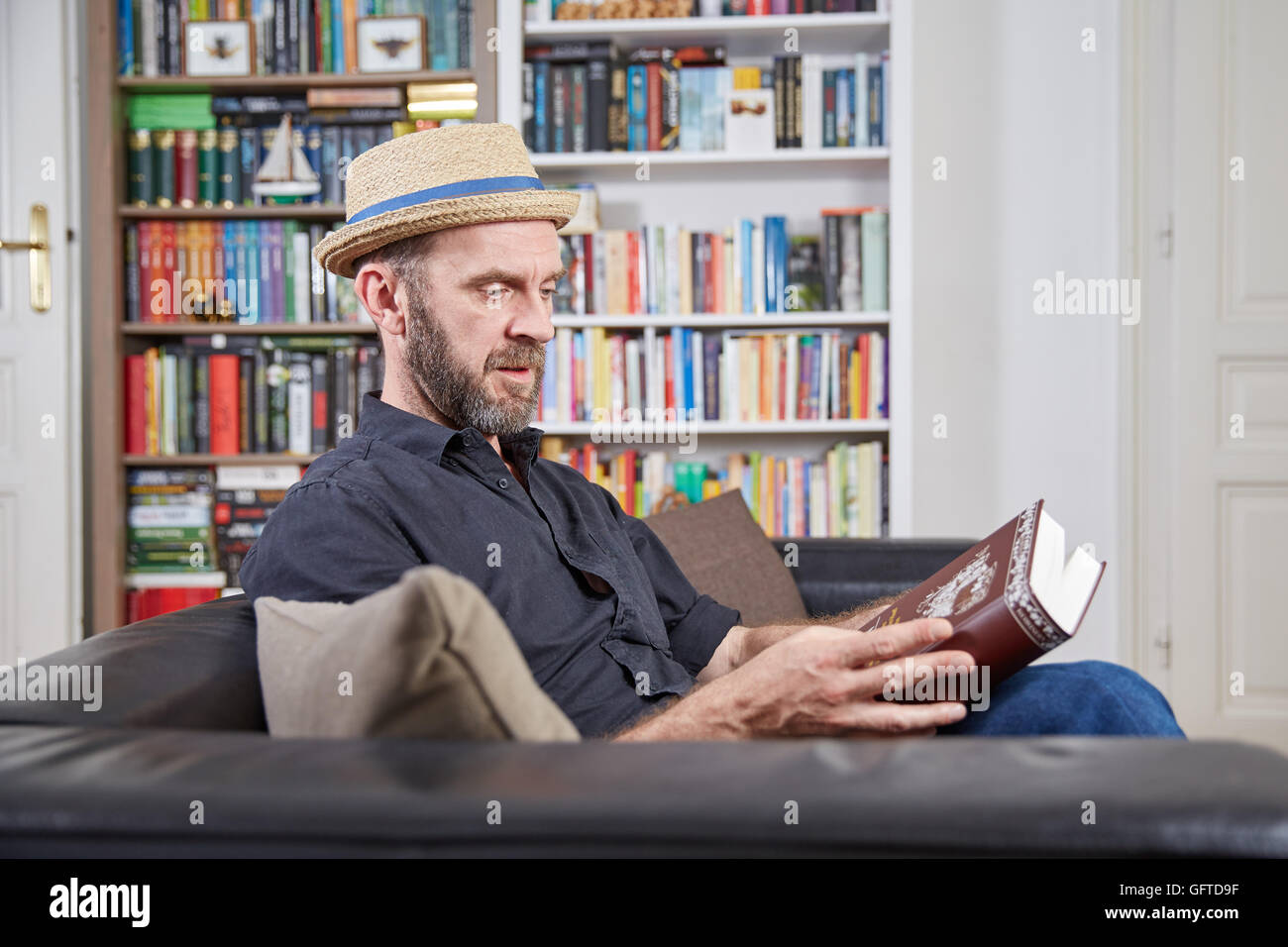 Student mit einem Buch vor einem Bücherregal auf einer Couch Hut Stockfoto