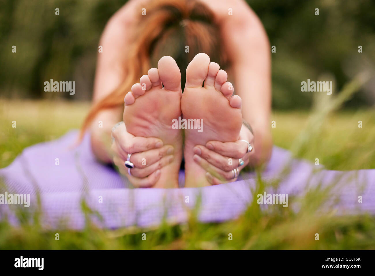Fitness-Frau, die auf der Trainingsmatte sitzt, sich nach vorne beugt und die Füße hält. Frau übt Paschimottanasana Yoga auf Gras. Fokus Stockfoto