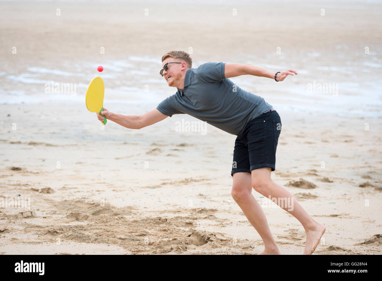 Ein junger Erwachsener Mann spielen mit einem Schläger und Ball an einem Strand, stretching, um den Ball zu erreichen Stockfoto