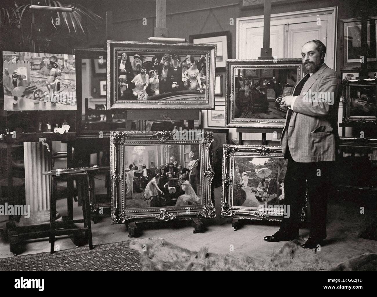 Französischer Maler und Karikaturist Albert Guillaume in seinem Atelier in Paris. Er posiert vor seiner berühmten Werke: "Nachzügler (oben in der Mitte) und"The Lie"(oben rechts). Albumen print c.1914 Stockfoto