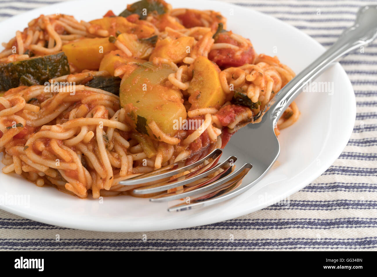 Angel Hair Pasta mit Zucchini und Spinat auf einem weißen Teller mit einer Gabel oben auf eine gestreifte Tischdecke. Stockfoto