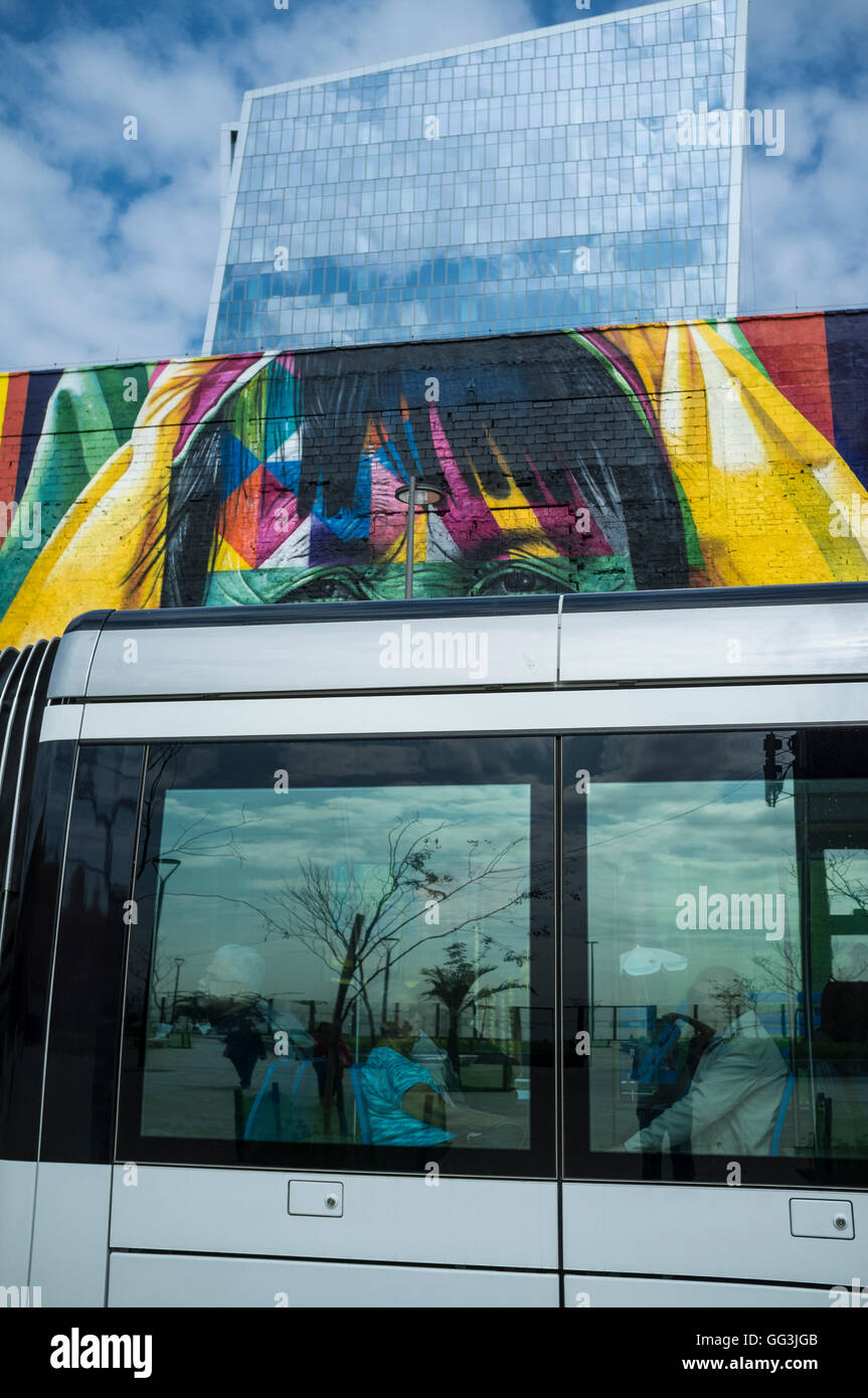 Rio De Janeiro Light Rail (Portugiesisch - VLT do Rio De Janeiro oder VLT Carioca), eine Stadtbahn eröffnet im Juni 2016 vor 2016 Olympischen Spiele Pässe vor Eduardo Kobra s Wandbild benannte Ureinwohner aus 5 Kontinenten (Povos Nativos Dos 5 Continentes) am Boulevard do Porto in Rio De Janeiro Hafen umgebenden Bereich, Brasilien, Teil des Projekts Porto Maravilha (wunderbare Hafen Programm) , ein Revitalisierungsprojekt der Stadt Port Zone. Kobra ist eine brasilianische Street-Artist bekannt für große Wandbilder, Gemälde in der Regel mit Porträts mit einer Technik der Wiederholung Quadrate und trian Stockfoto