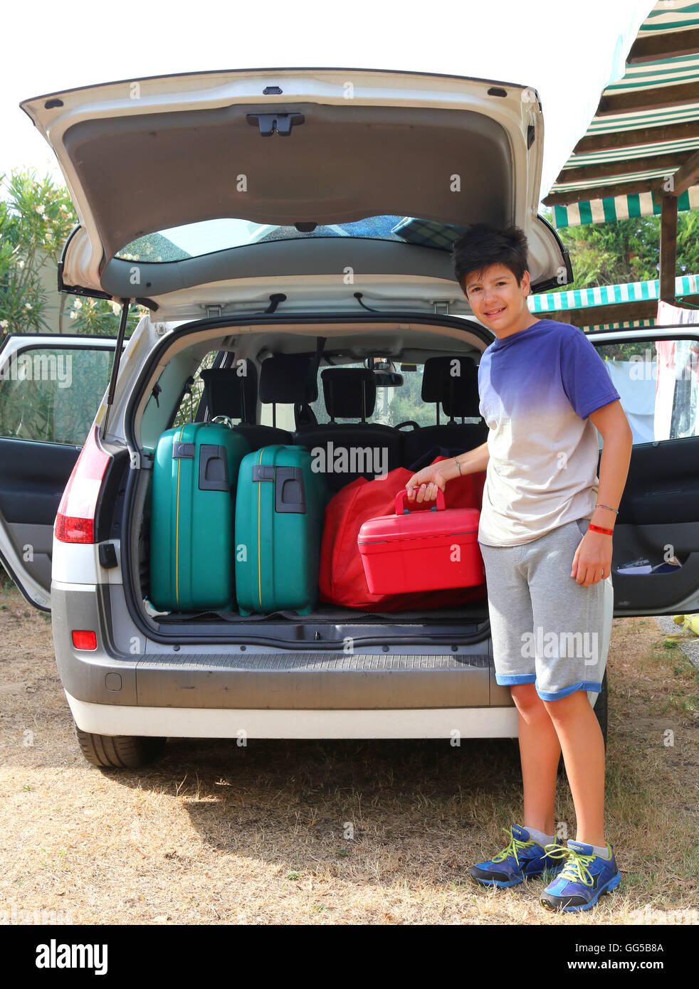 kleiner Junge lädt einen wenig roten Koffer in den Kofferraum des Autos vor seiner Abreise nach einer langen Reise Stockfoto