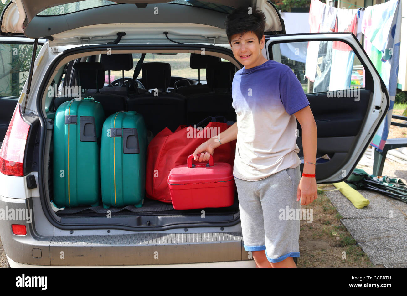 lächelnde junge lädt einen wenig roten Koffer in den Kofferraum des Autos vor seiner Abreise nach einer langen Reise Stockfoto