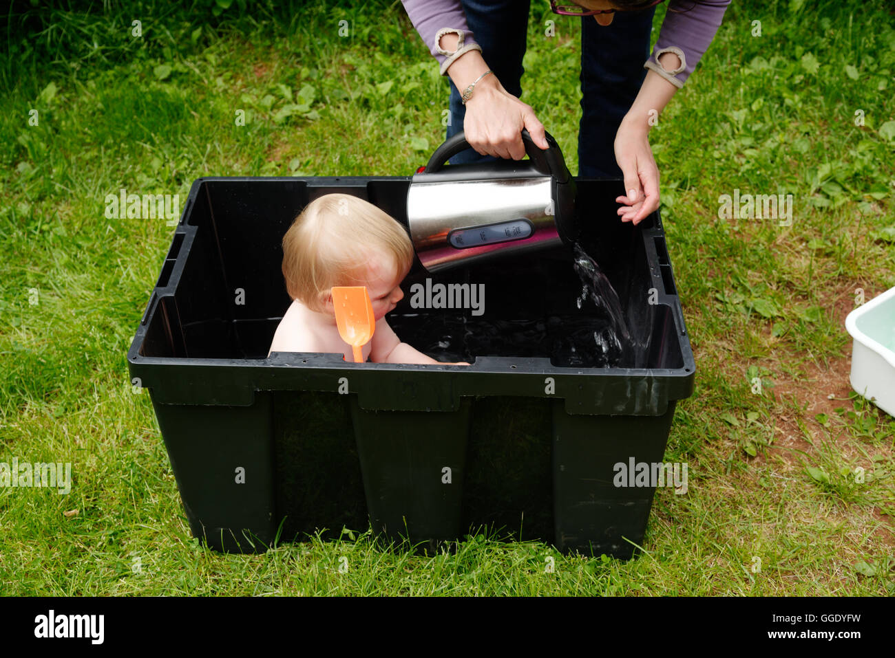 Ein kleines Mädchen (2 Jahre alt) in einer Kunststoffbox gebadet, während Camping, Mom fügt warmes Wasser aus einem Wasserkocher Stockfoto