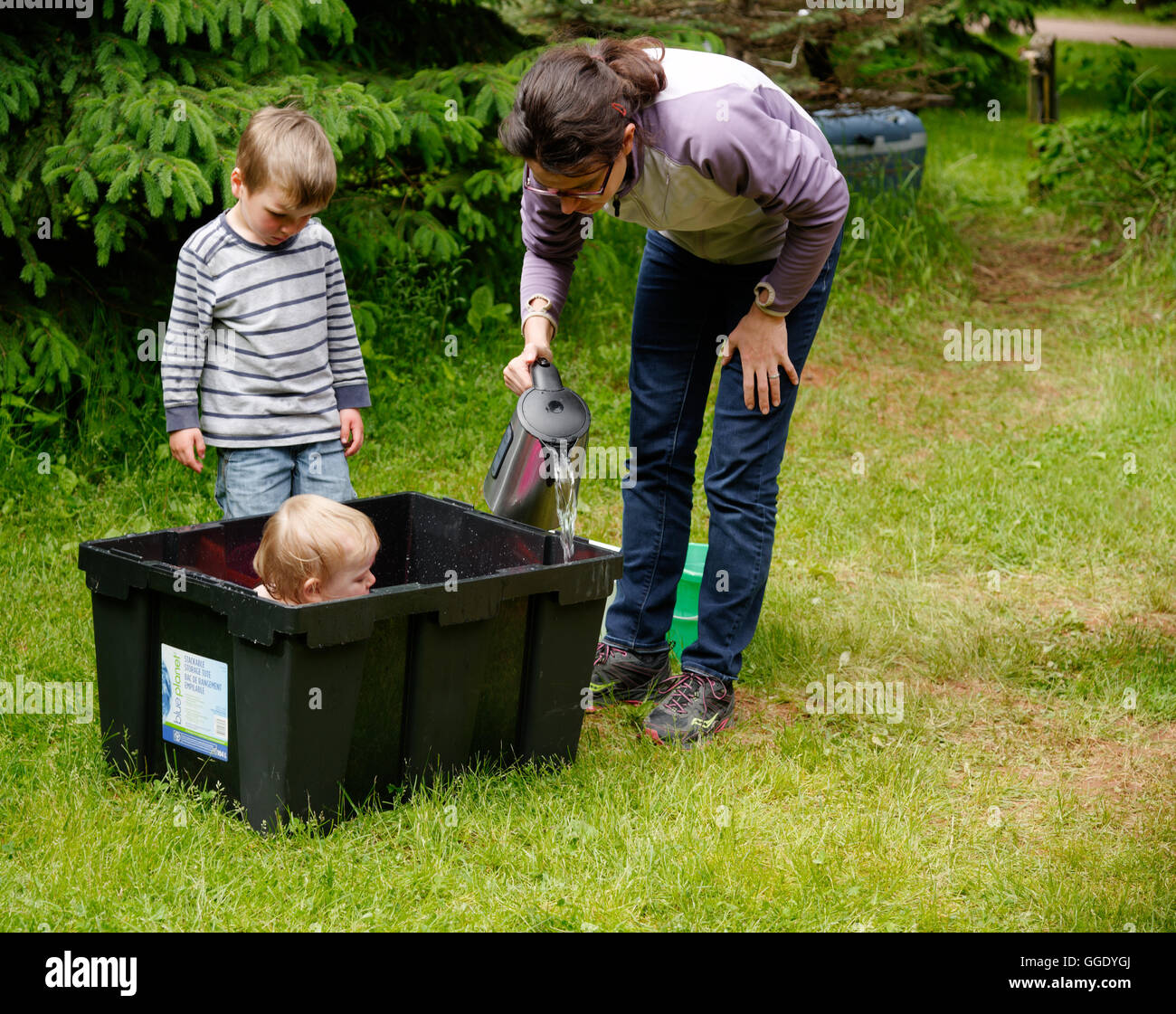 Ein kleines Mädchen (2 Jahre alt) wird in einer Plastikbox gebadet, während camping Mama warmes Wasser aus einem Wasserkocher und Bruder Uhren fügt Stockfoto