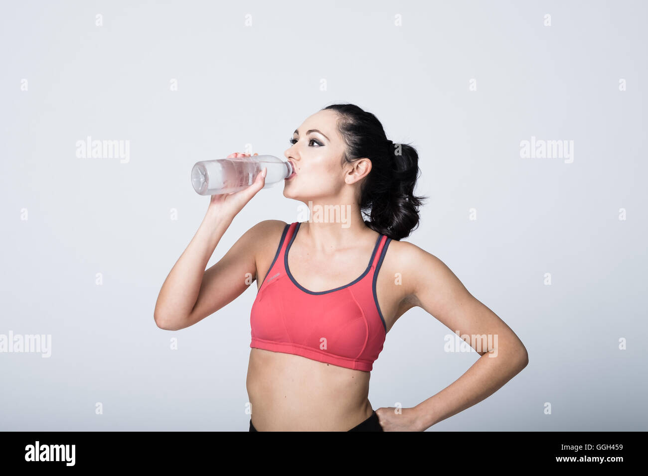 Fitte Frau in ihren 20ern, die nach einem Workout Wasser trinkt. Sie trägt pinkfarbene Trainingsbekleidung vor weißem Hintergrund. Kopieren Sie den Platz nach links Stockfoto