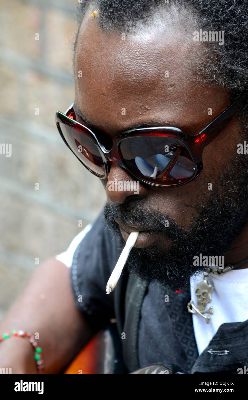 Schwarzen Straßenmusiker mit Sonnenbrille und Zigarette & Schädel Anhänger London England Stockfoto