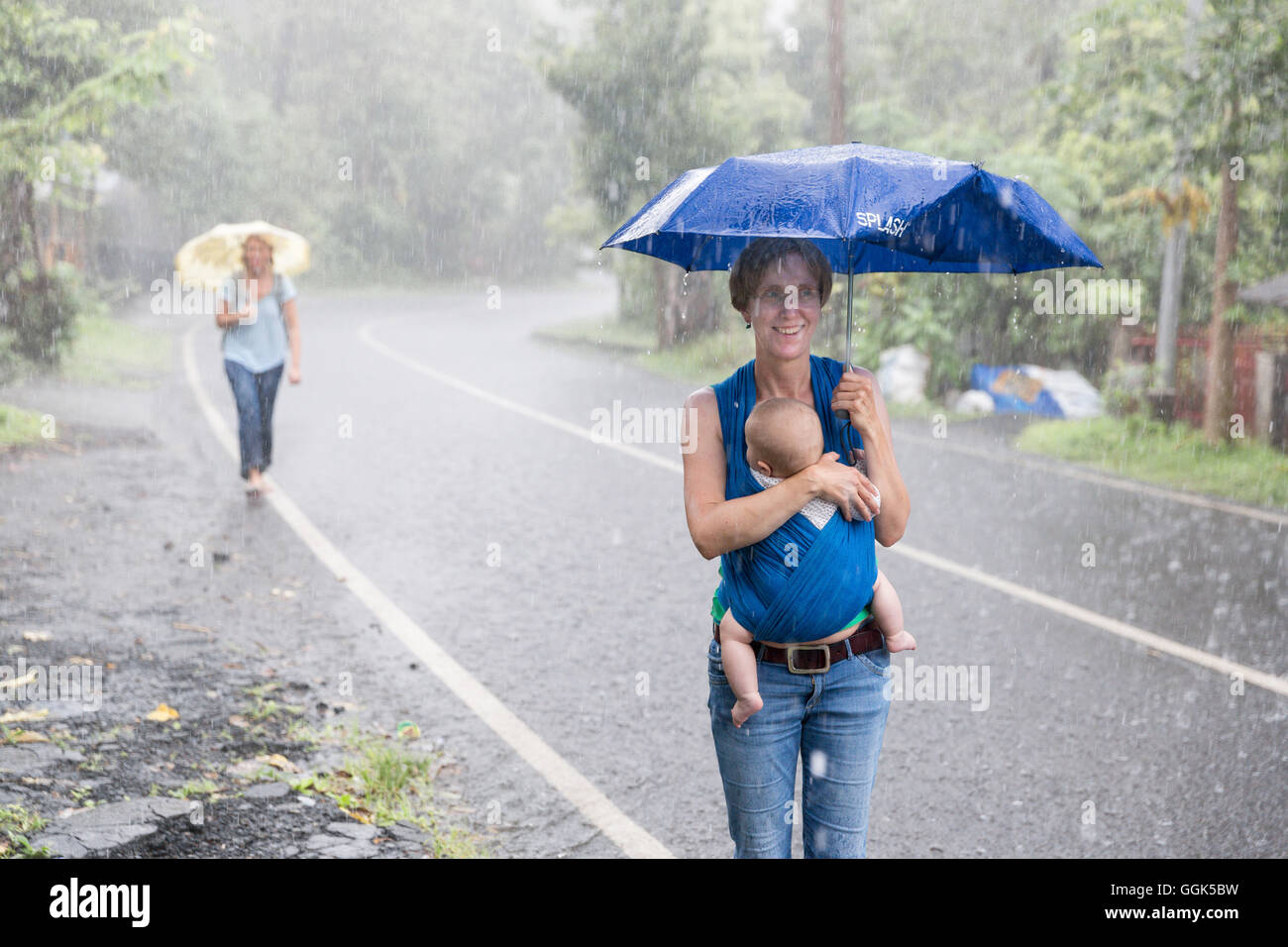 Tropenregen, Monsun, Starkregen, Touristen zu Fuß entlang einer Straße, deutsche Mutter mit ihrem Baby, wickeln, Baby-Tragetuch, 5 Monate ol Stockfoto