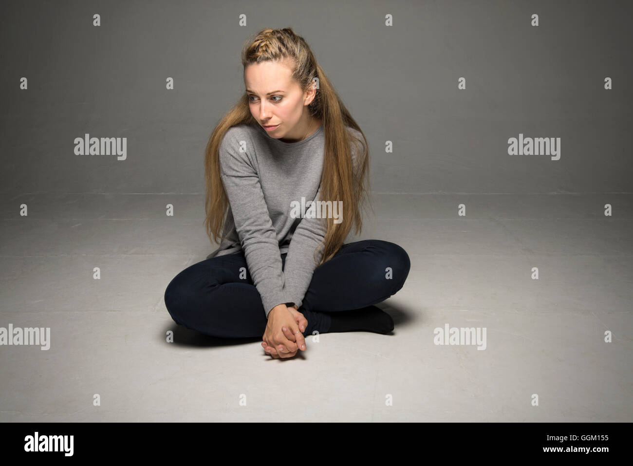 Traurige junge Frau sitzt im leeren Raum am Boden gekreuzten Beinen fest umklammert vor weit Wegschauen am Boden Stockfoto