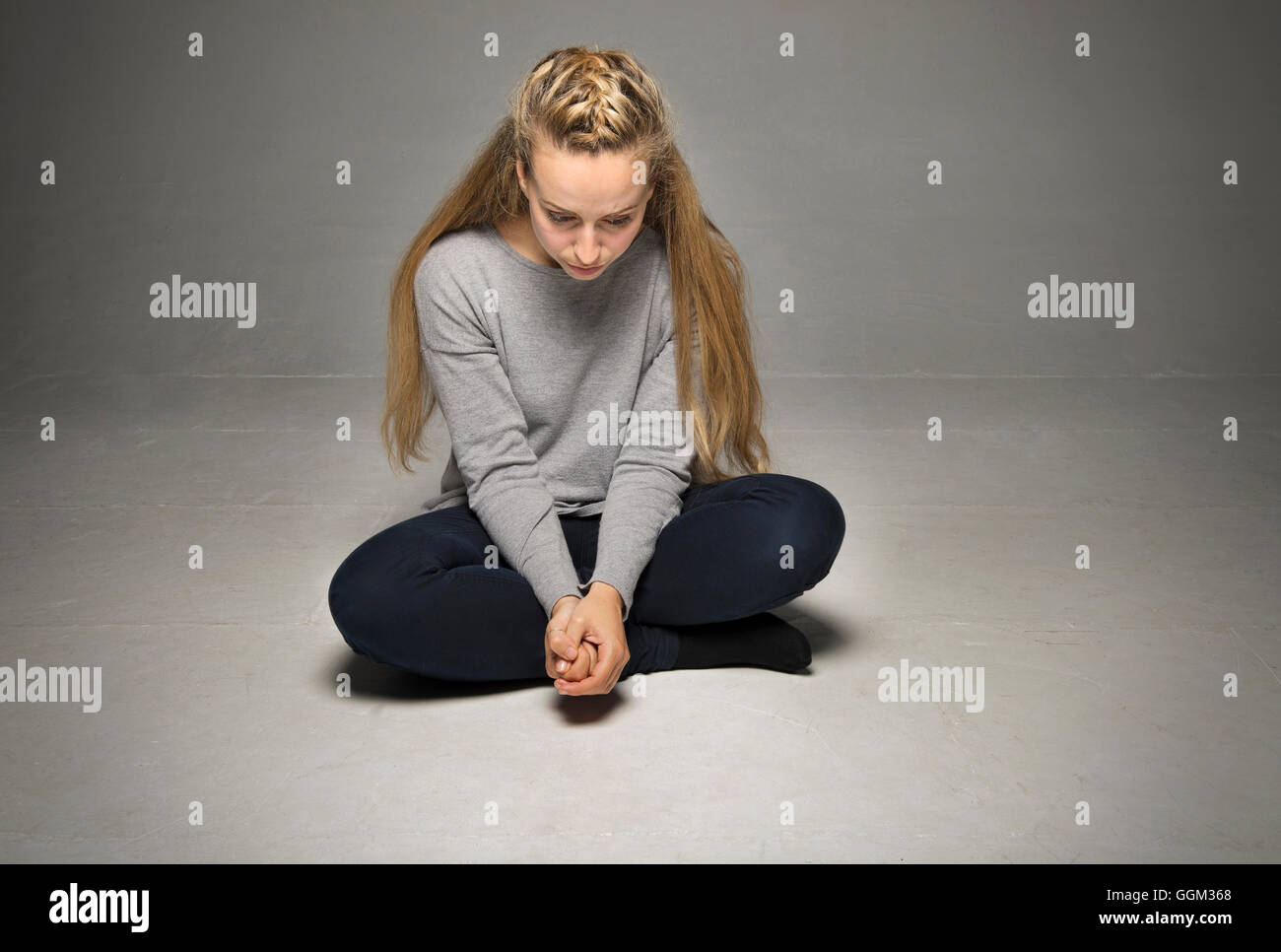 Traurige junge Frau sitzt im leeren Raum am Boden Beine gekreuzt Händen fest umklammert vorne nach unten Stockfoto