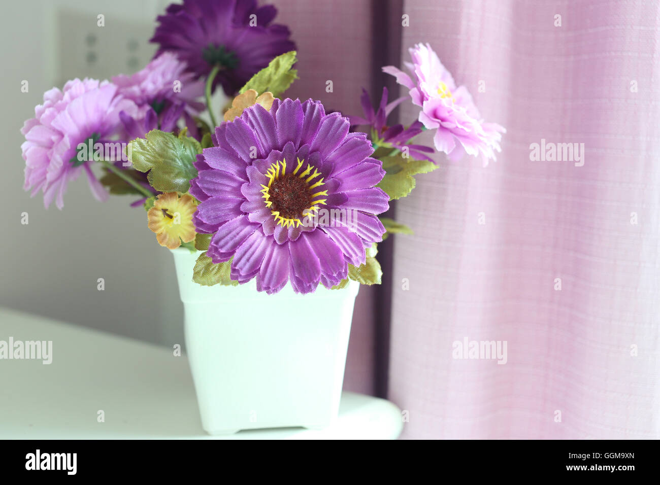 Künstliche lila Blüten in einer weißen Vase platziert neben dem Fenster im Schlafzimmer. Stockfoto