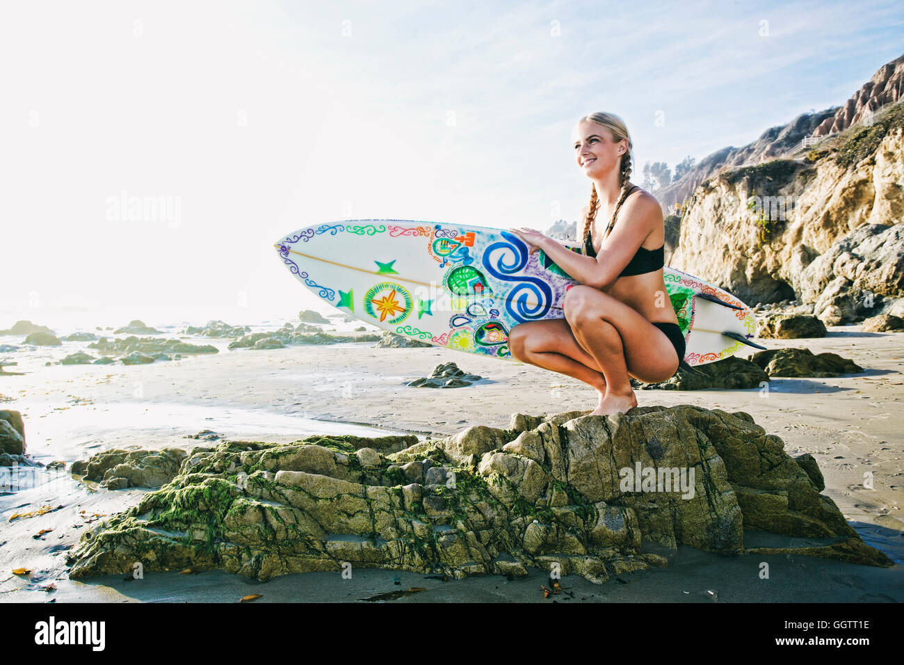 Kaukasische Frau hocken auf Felsen mit Surfbrett am Strand Stockfoto