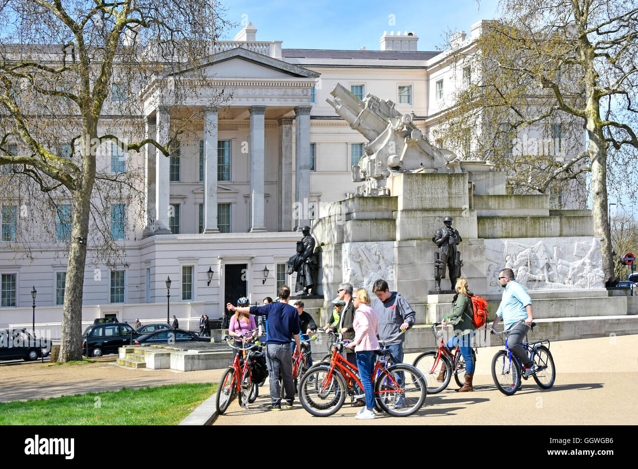 Tourguide (zeigend) mit Zyklus Reisegruppe neben dem The Royal Artillery-Denkmal am Hyde Park Corner London UK mit Lanesborough Hotel darüber hinaus Stockfoto