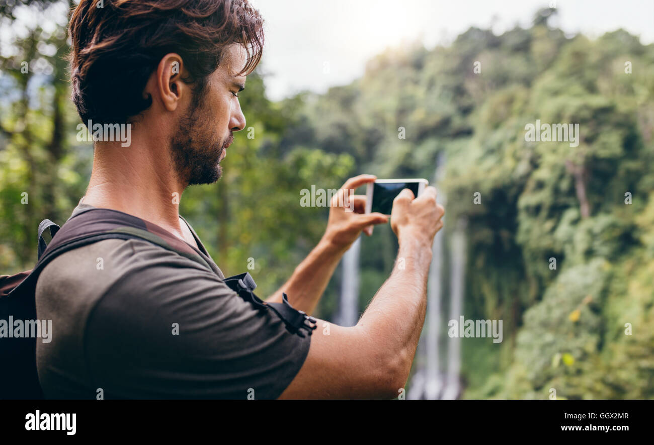 Mann mit Rucksack stand vor Wasserfall und Aufnehmen eines Fotos mit seinem Handy. Männliche Wanderer fotografieren einen Wasserfall in Stockfoto