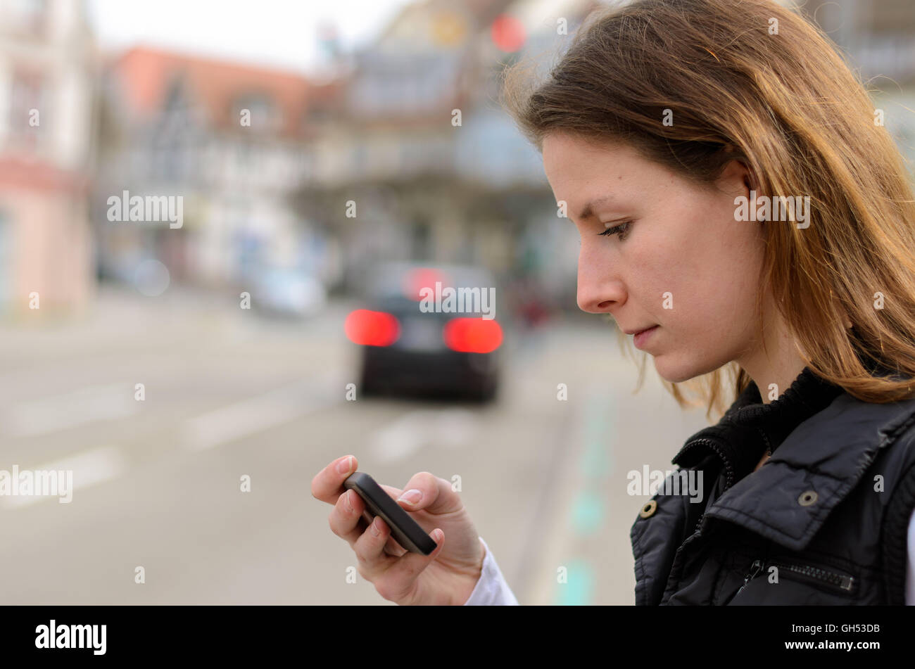 Junge Frau, die SMS-Nachrichten auf ihr Handy gesucht, wie sie wartet ein die Straße an einem roten Fußgängerüberweg überqueren Stockfoto