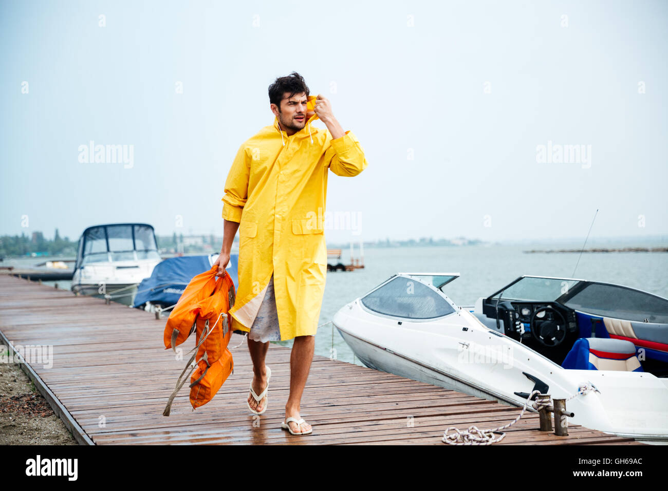 Junge hübsche Seemann Mann in gelben Mantel zu Fuß am Meer Pier holding Schwimmweste Stockfoto