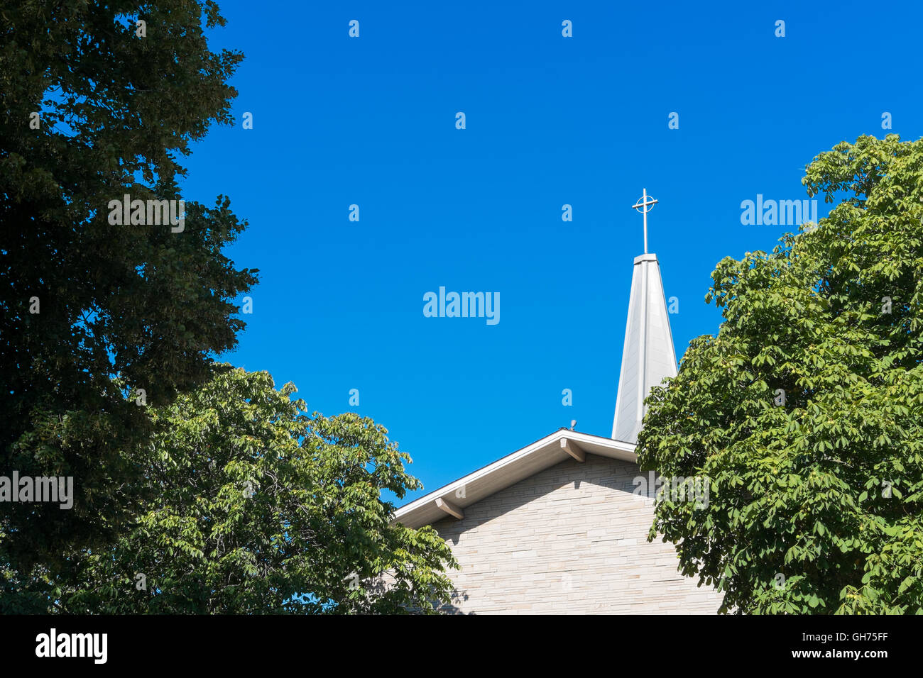 Ein Kirchturm mit einem Kreuz etwas verdeckt durch Bäume mit einem blauen Himmel an einem sonnigen Tag Stockfoto