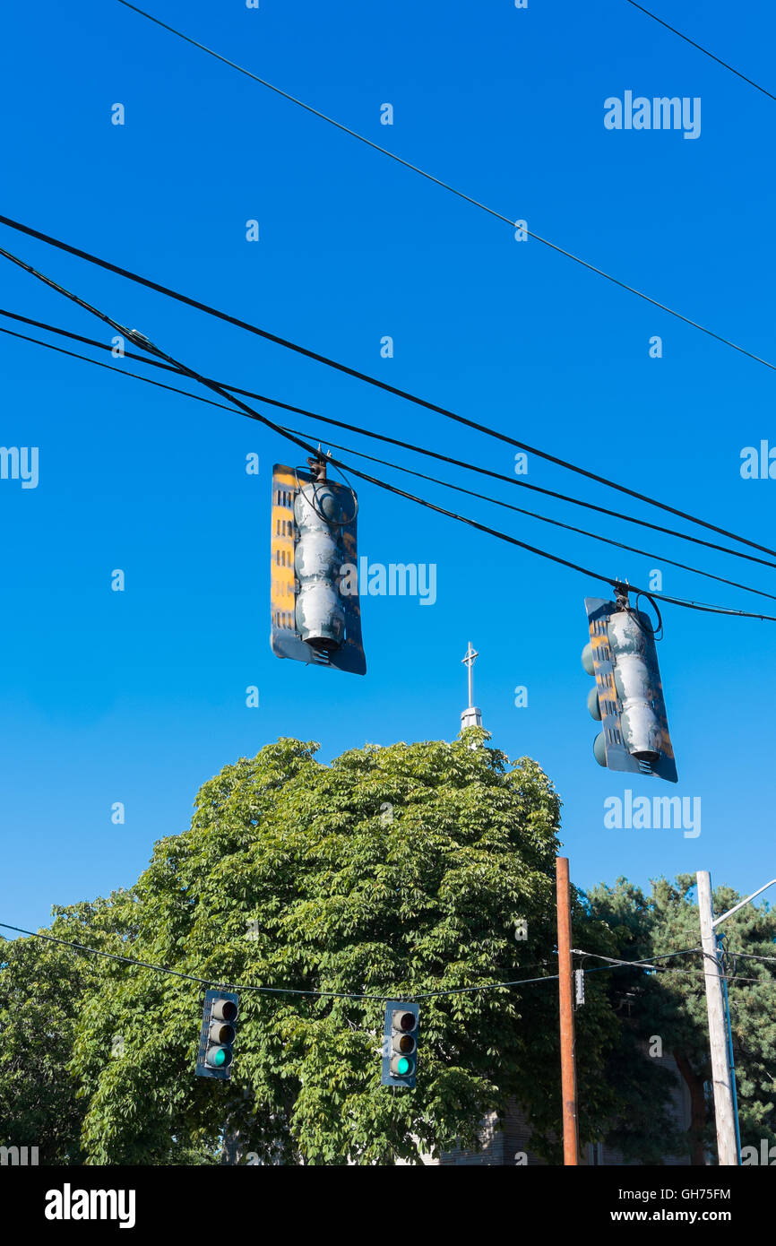 Ampel in der Kreuzung und ein Kirchturm mit einem Kreuz etwas verdeckt durch Bäume mit einem blauen Himmel Stockfoto