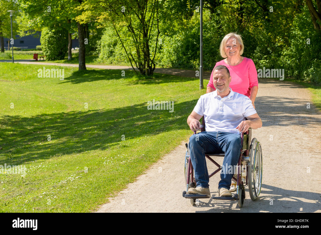 Mann im Rollstuhl sitzt, während Frau trägt rosa Bluse ihn weg Park an einem sonnigen Nachmittag schiebt Stockfoto
