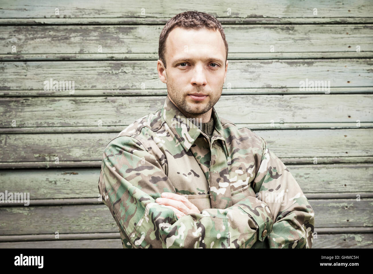 Ernsthafte kaukasischen militärischen Jüngling in Camouflage uniform mit Händen überqueren. Closeup Portrait über grüne Holzwand Stockfoto