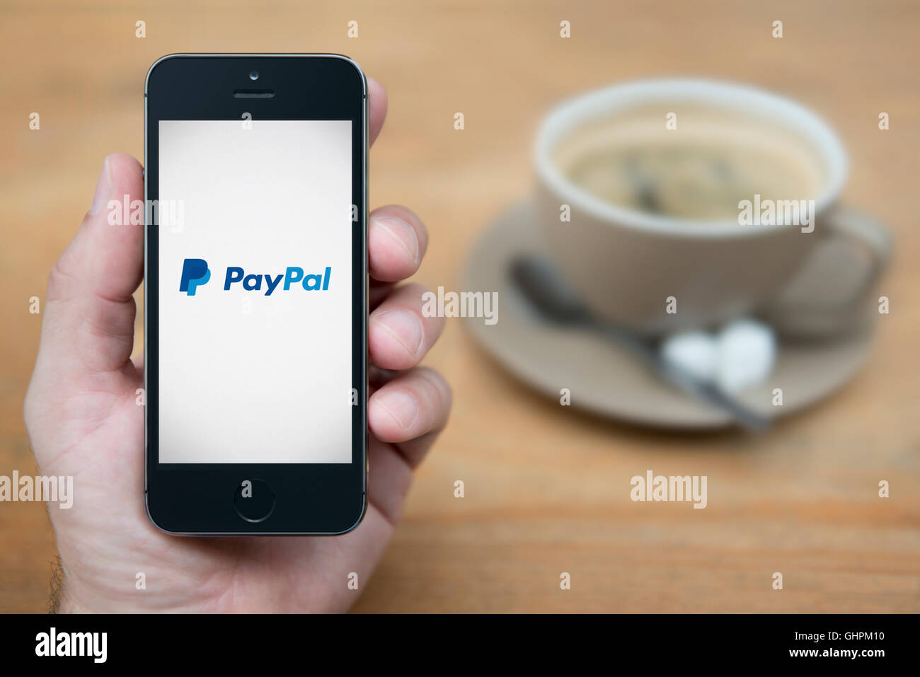 Ein Mann schaut auf seinem iPhone das das PayPal-Logo anzeigt, während bei einer Tasse Kaffee (nur zur redaktionellen Verwendung) saß. Stockfoto