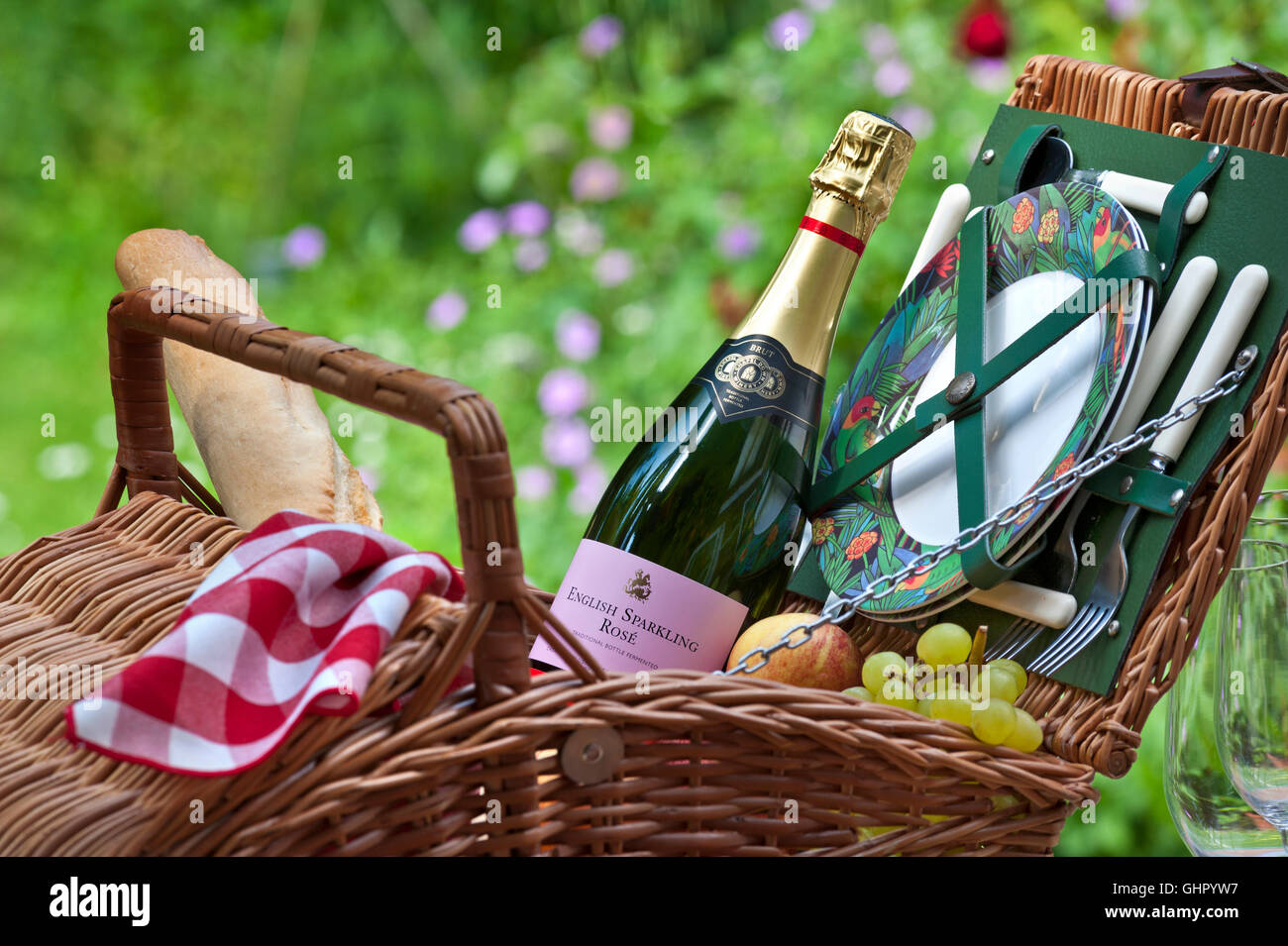 Englischer Sparkling Rosé Weinflasche Weidengeflecht Picknickkorb Korb Korb mit Baguette Trauben Obst etc. Sonniger Frühling Sommer im Freien florale britische Gartensituation Stockfoto