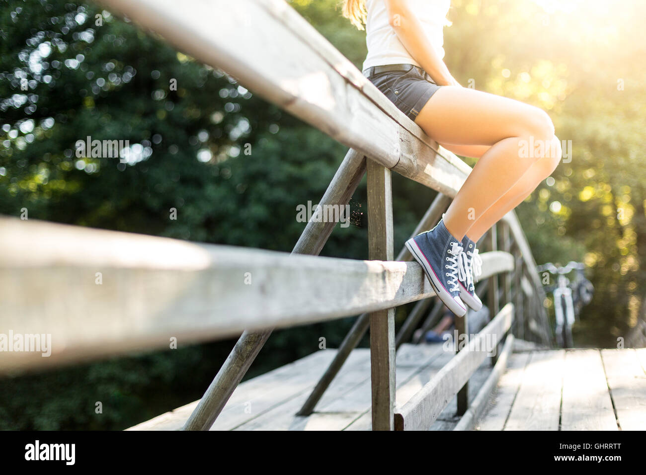Junge Frau mit schönen sportlichen Beinen sitzen auf einer Holzbrücke Geländer in Jeans Turnschuhen Stockfoto